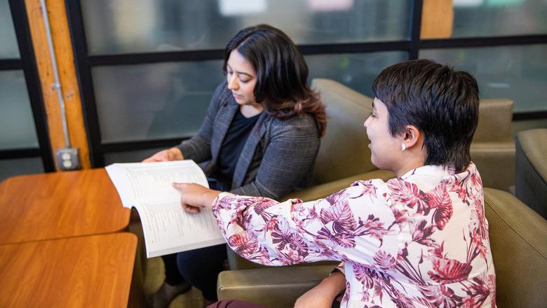 Two people sit next to each other in an office setting. One is holding a sheet of paper or binder while the other points to it.